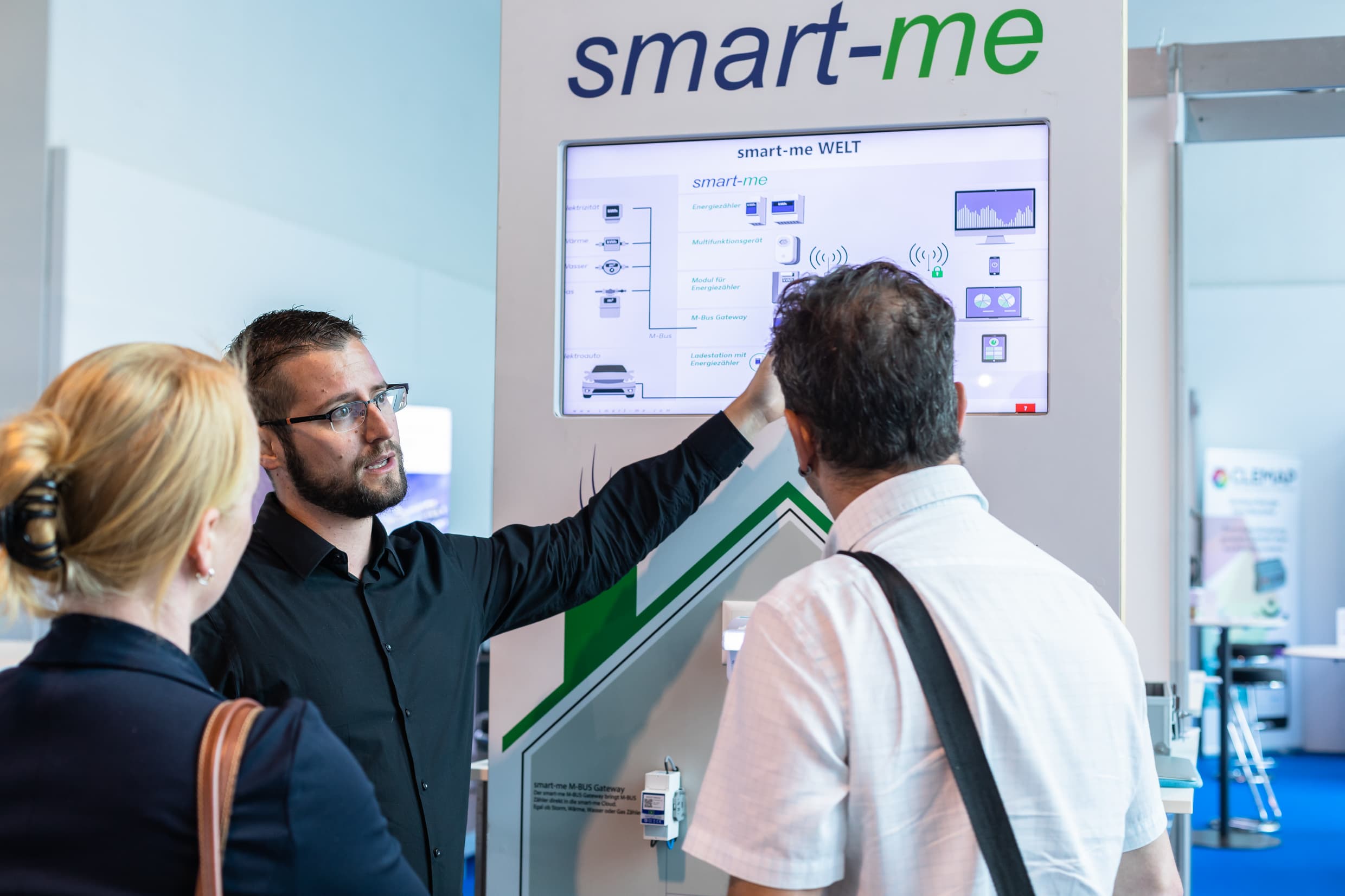 An exhibitor explains something on the screen to two interested visitors, smart-me.